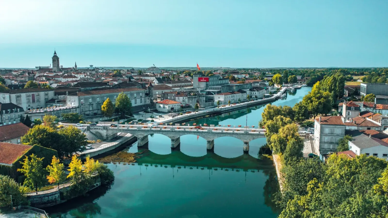 Vue aérienne d'une ville avec un pont sur une rivière, des bâtiments historiques, des toits rouges, un logo Heineken et des arbres verdoyants.