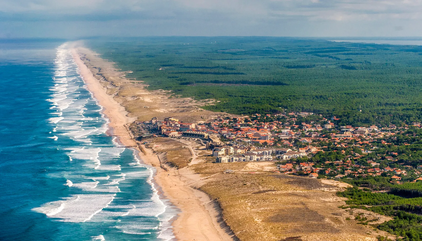 Vue aérienne d'une côte avec vagues blanches, plage de sable, petite ville aux toits rouges, entourée de forêt dense sous un ciel nuageux.