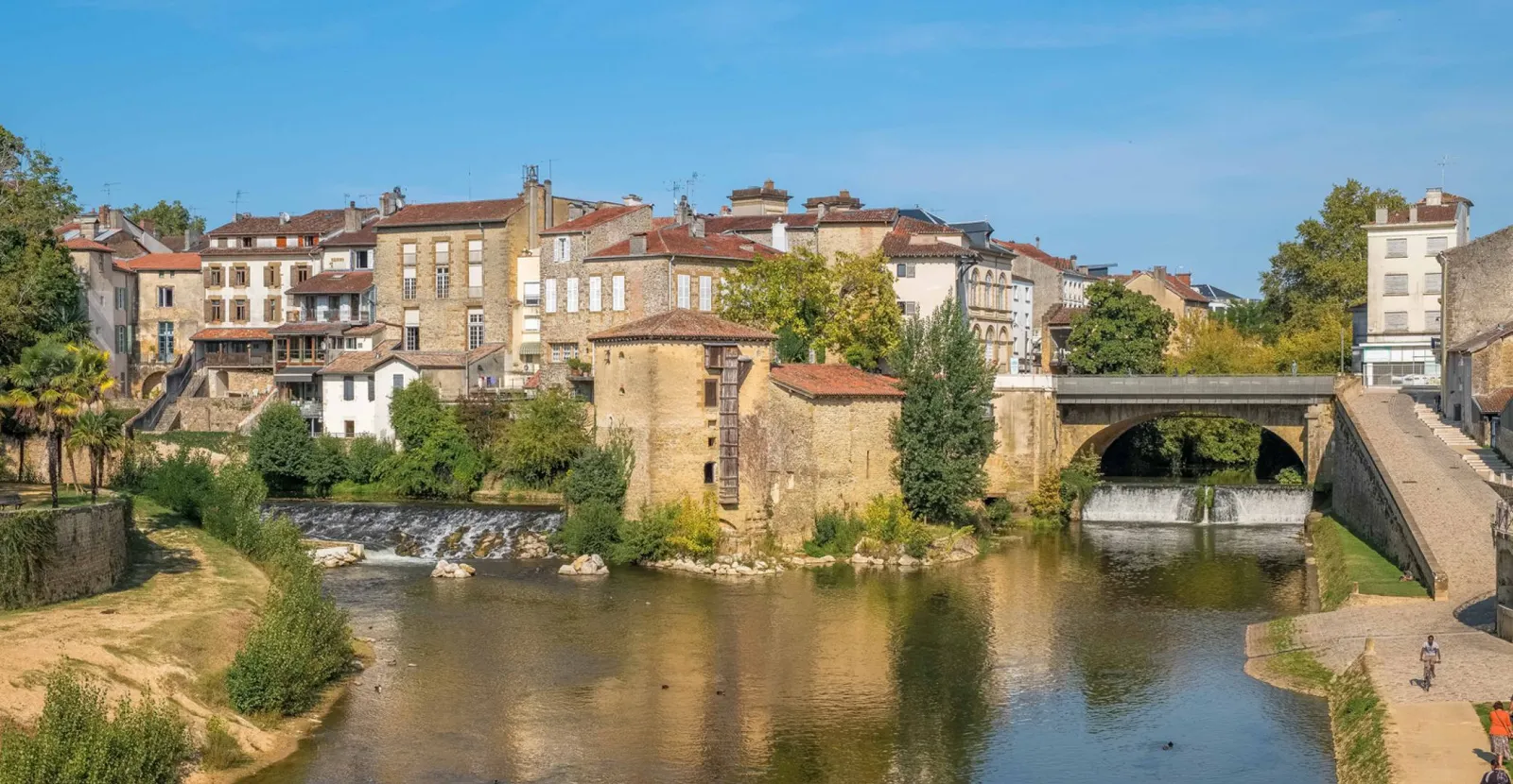 Ville pittoresque avec maisons en pierre près d'une rivière calme et pont. Ciel bleu, végétation luxuriante, ambiance sereine.
