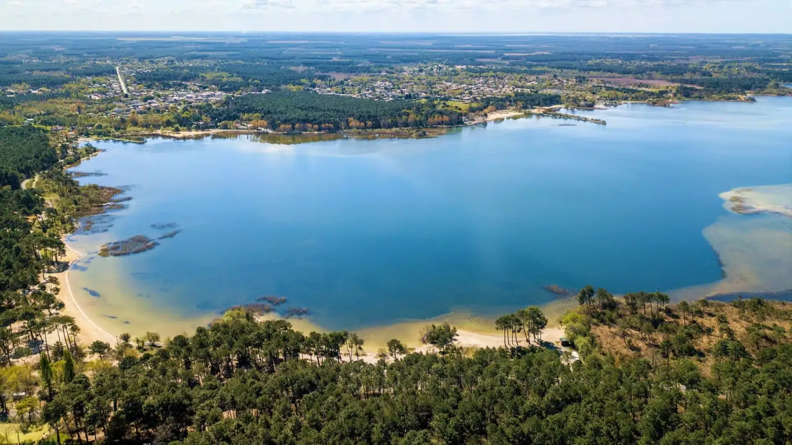 Vue aérienne du lac de Sanguinet entouré de forêts vertes et d'une ville au loin sous un ciel clair. Atmosphère paisible et naturelle.