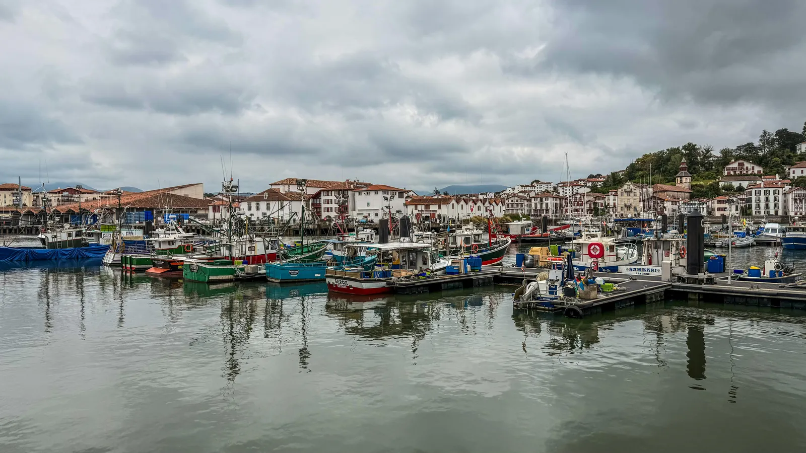 Port avec bateaux colorés amarrés, arrière-plan de bâtiments blancs au toit rouge, ciel nuageux. Atmosphère calme et paisible.
