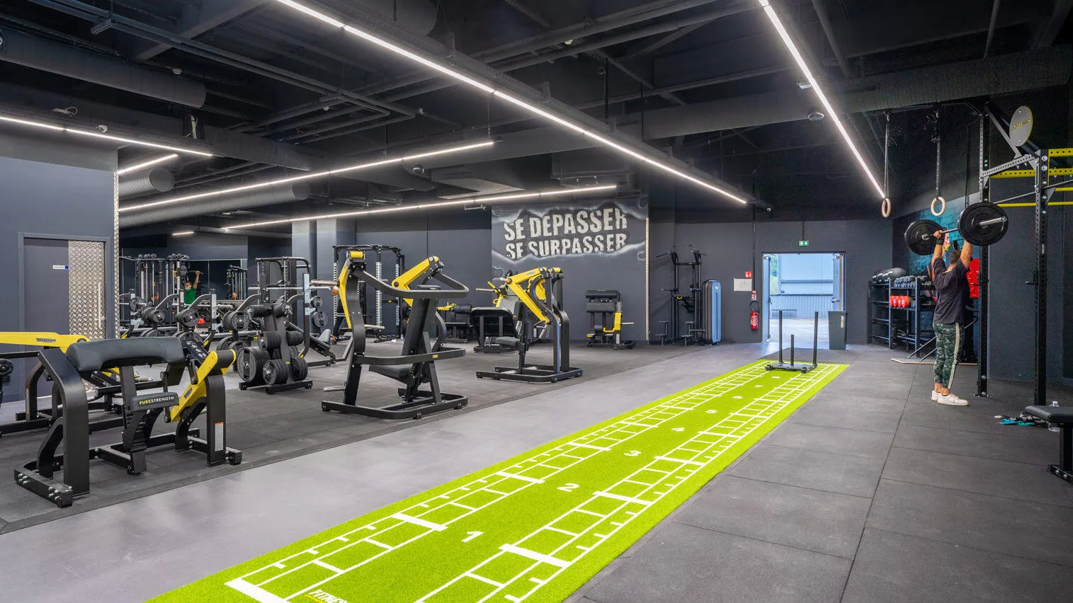 Gym moderne avec équipements noirs et jaunes. Personne soulevant des haltères. Piste verte au sol. "SE DEPASSER, SE SURPASSER" au mur.