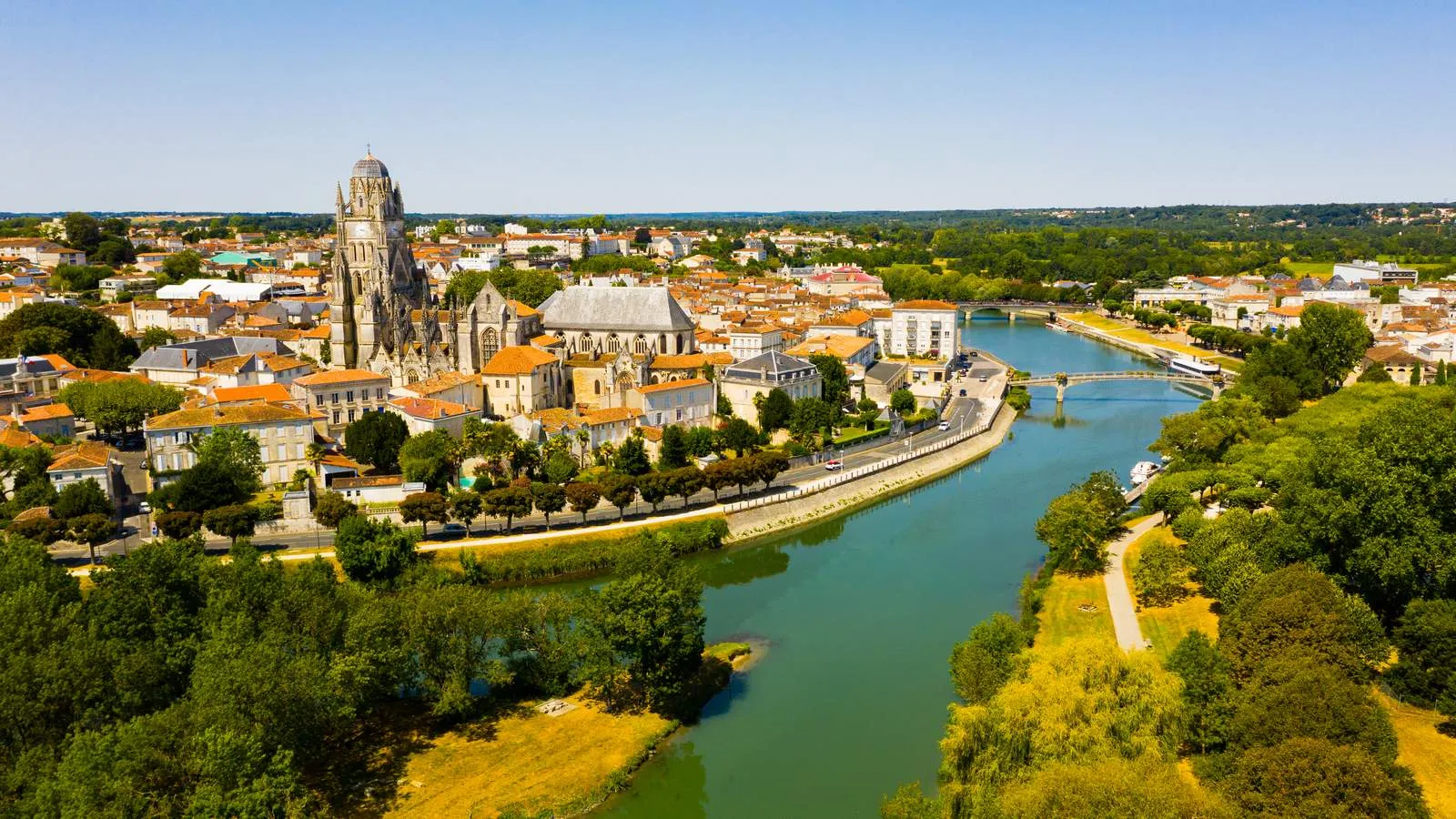Vue aérienne d'une ville avec une cathédrale gothique, toits orange, rivière sinueuse, pont. Végétation verdoyante et ciel bleu clair.